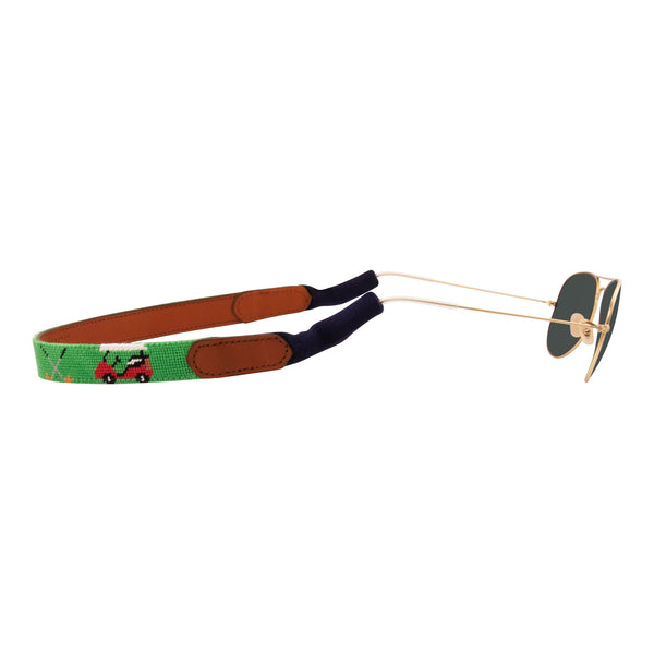 Side view golfing theme handmade needlepoint sunglass strap showing golf carts golf clubs golf balls against a green background with sturdy cotton covered silicone ear connectors and a soft leather backing