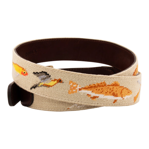 Side view of Sportsman Needlepoint Belt with khaki colored background showing outdoor sporting themed images of a pointer dog, crossed shotguns, shotgun shell, fly fishing rod, redfish, fishing boat, flying duck and dove.  Belt has brass buckle and full-grain leather backing.