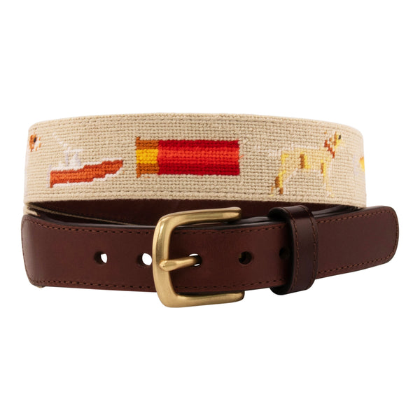 Front view of Sportsman Needlepoint Belt with khaki colored background showing outdoor sporting themed images of a pointer dog, crossed shotguns, shotgun shell, fly fishing rod, redfish, fishing boat, flying duck and dove.  Belt has brass buckle and full-grain leather backing.