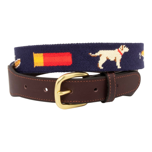 Front view of Sportsman Needlepoint Belt with navy blue background showing outdoor sporting themed images of a pointer dog, crossed shotguns, shotgun shell, fly fishing rod, redfish, fishing boat, flying duck and dove.  Belt has brass buckle and full-grain leather backing.